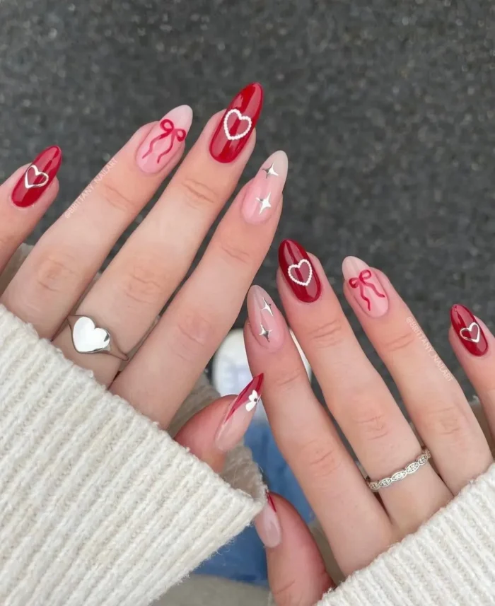A detailed romantic red nail design on almond nails, featuring a mix of hearts, bows, and sparkle accents.