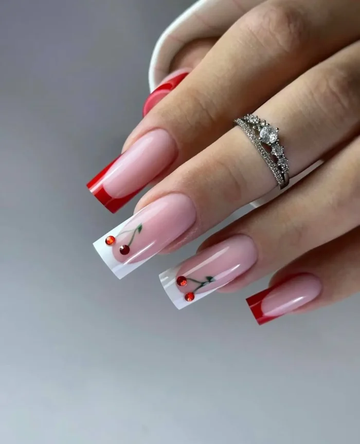 Long square nails featuring a playful combination of classic red French tips and white French tips adorned with cute cherry designs made from red rhinestones and painted green stems.