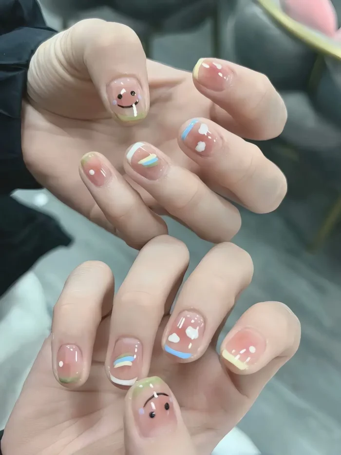 Very short rounded nails with a sheer base featuring cute weather-themed art: smiley faces, clouds, rainbows, and pastel French tip variations.