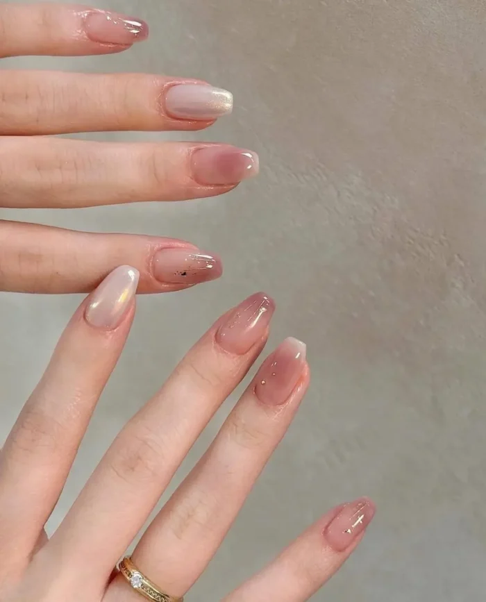Almond nails with sheer rosy nude bases, subtle shimmer effects, tiny gold flakes, and small gold cross shape accent.