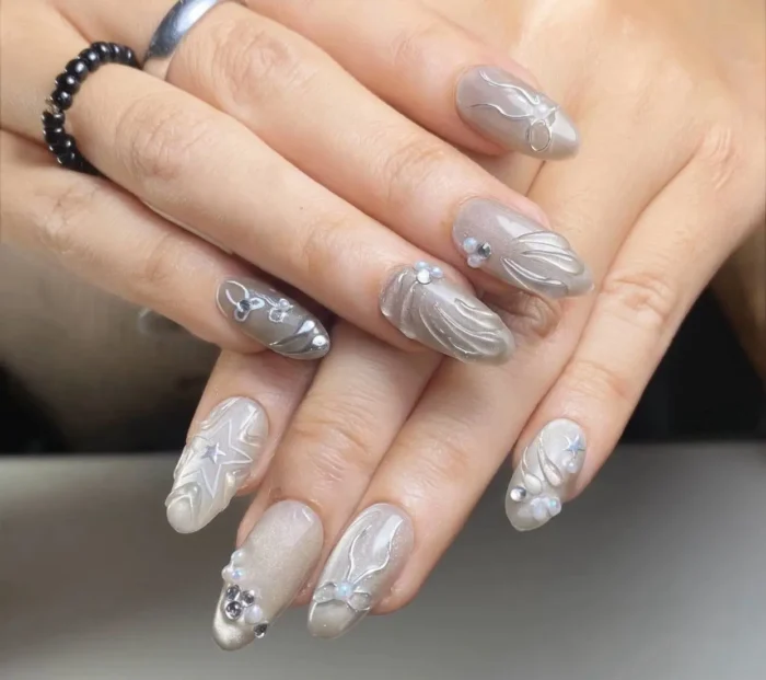 Almond nails with a pearlescent grey base featuring intricate dimensional silver designs like swirls, stars, and bows, accented with rhinestones.