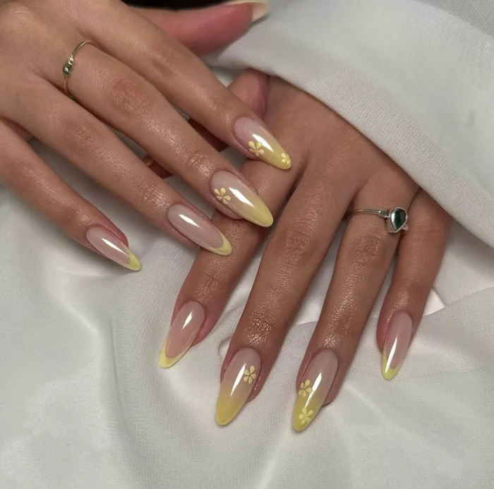 Long almond shaped nails with sheer nude base and pearlescent pastel yellow French tips, accented with small simple floral designs.