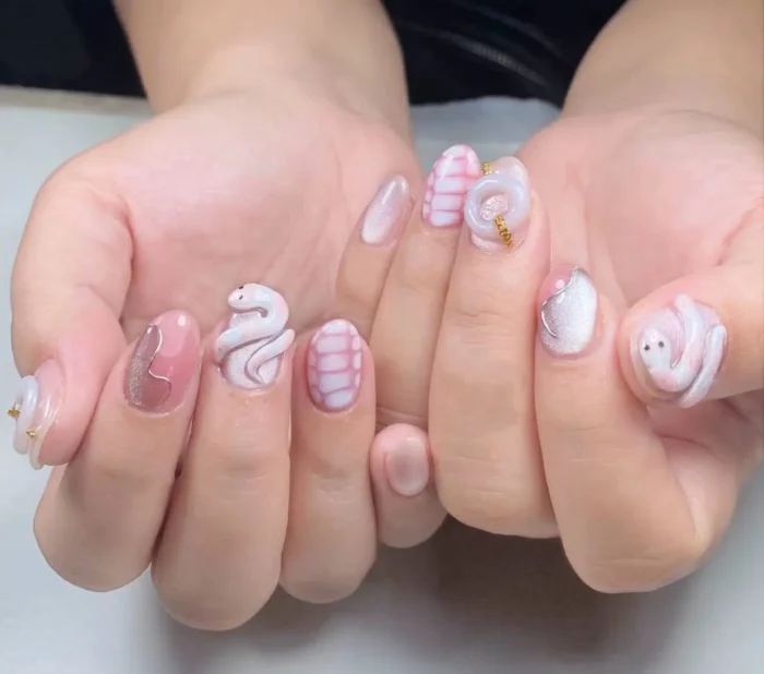Oval nails with mixed designs including 3D sculpted snakes and ghosts, pink plaid patterns, silver cat-eye effect, and dusty rose polish.