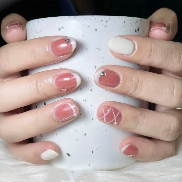 Short rounded nails with dusty rose and off-white polish featuring white geometric line art, double-ended curves, and small rhinestone accents.
