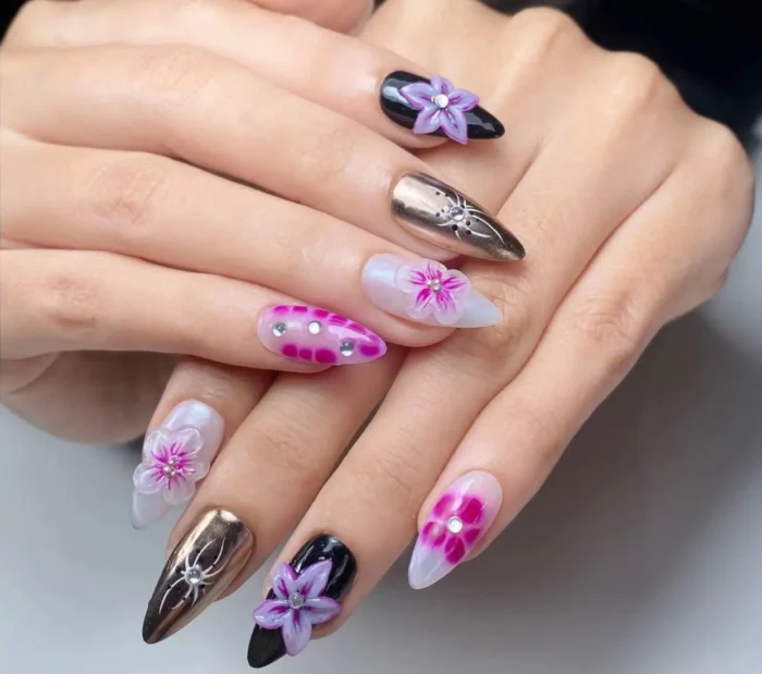 Long stiletto nails with mixed designs including black with 3D lavender flowers, metallic bronze chrome with rhinestones, and pink checked patterns.