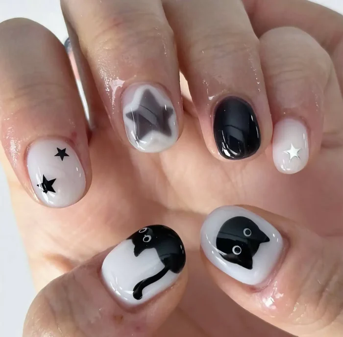 Short rounded nails with a black and white theme featuring black cat illustrations, painted and dimensional stars, and silver star accents.