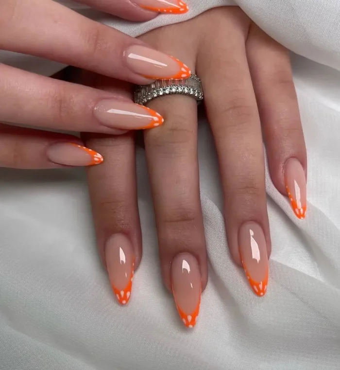 Long almond-shaped nails showcasing a vibrant neon orange V-shaped French tip, creatively accented with a pattern of small white dots.