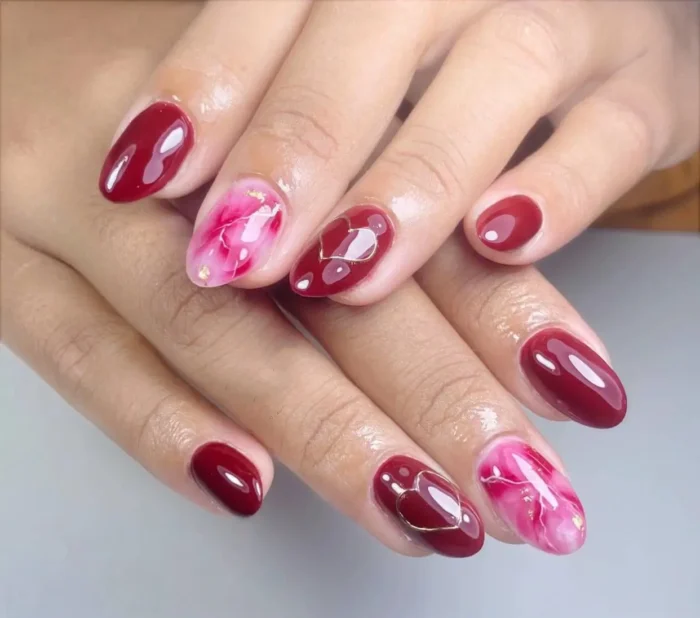 Oval nails featuring glossy ruby red polish, pink and white marble accent nails with gold flecks, and gold heart outline details.