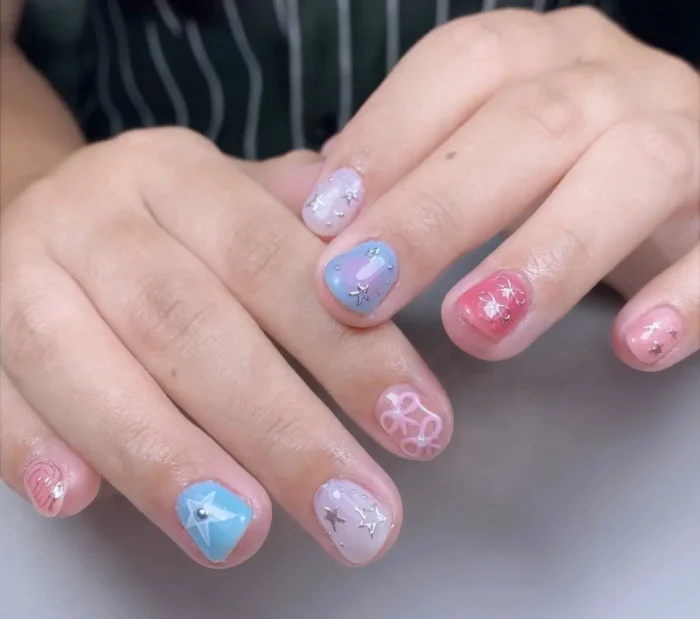 Short rounded nails with mixed pastel designs including baby blue, coral, and pearly lilac bases adorned with silver star charms, rhinestones, and white ribbon outlines.