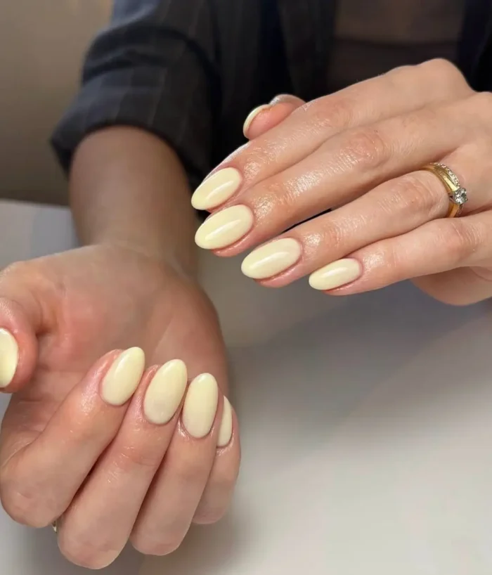A pair of hands showcasing a simple, elegant manicure with medium almond-shaped nails painted in a solid butter yellow color.