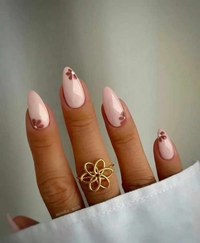 Soft pink almond nails adorned with small, minimalist brown flower designs near the tips.