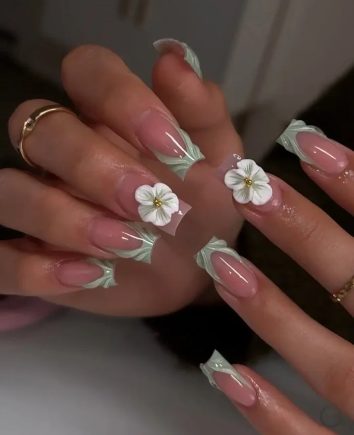 A detailed view of long sculpted nails with sage green architectural tips and white 3D flower accents with gold centers.