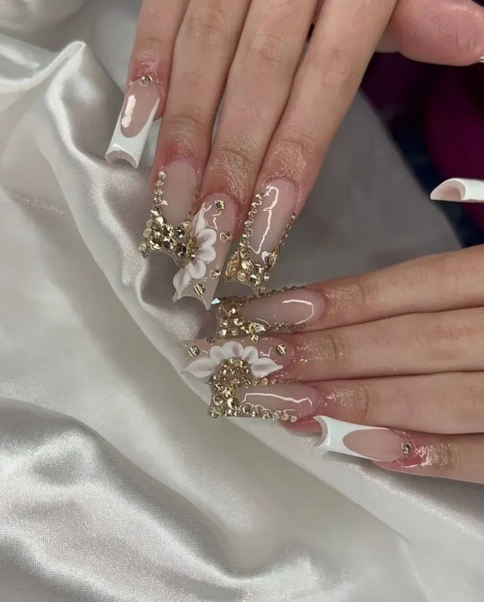 Long square nails with a white French manicure, extravagantly embellished with large white 3D flower charms and an abundance of gold crystal detailing.