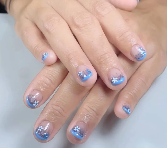 Short rounded nails with a sheer base, featuring sky-blue French tips covered in silver glitter and accented with small white daisy flowers.