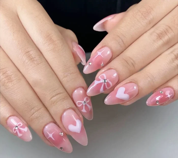 Almond nails with a sheer pink aura base featuring delicate white airbrushed bows, hearts, and sparkles, accented with rhinestones.