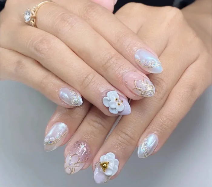Almond nails with a pearlescent white base featuring 3D white flowers, intricate gold wire details, and rhinestone and glitter accents.