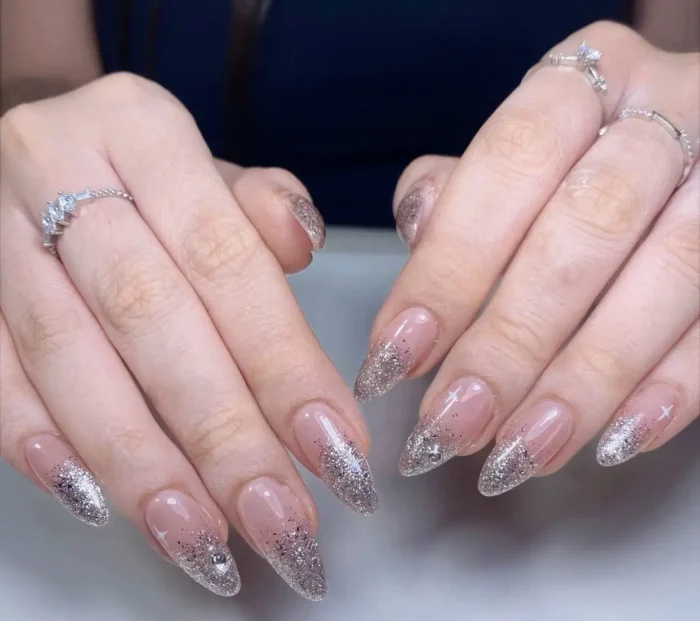 Long almond nails with a sheer nude base featuring a dense silver glitter gradient fading from the tips, with subtle starburst or rhinestone accents.