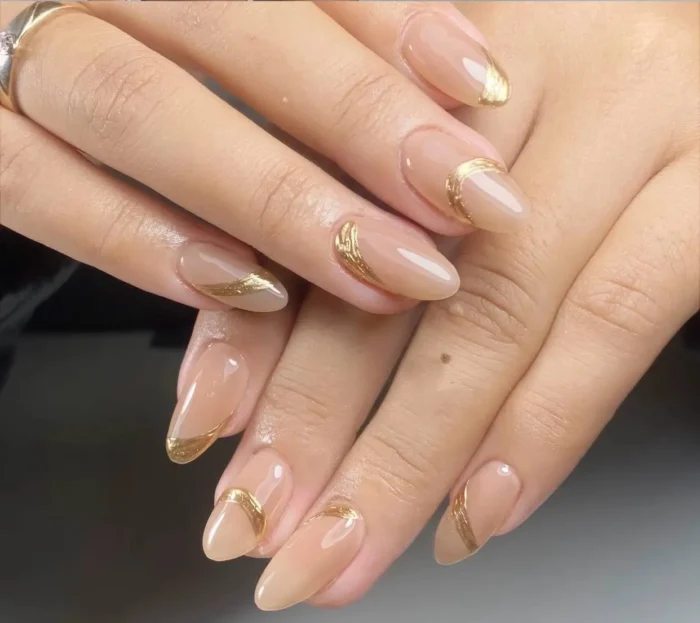 Almond nails with a sheer neutral beige base featuring abstract, dimensional gold metallic swoops and curved line accents.