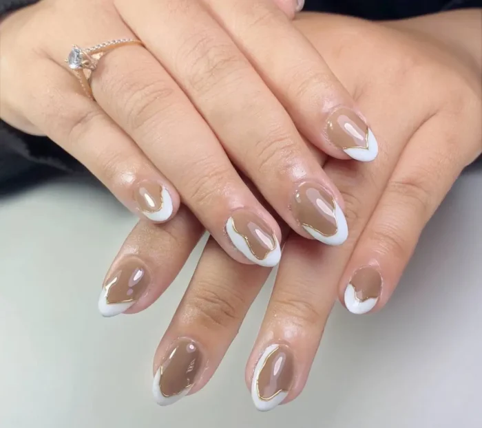 Almond nails with a negative space tip design featuring abstract taupe shapes outlined in white and then fine gold lines.