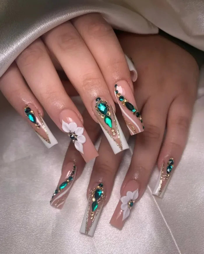 Long nude nails with white angled tips, 3D white flowers, emerald green rhinestones, and gold stripe details.