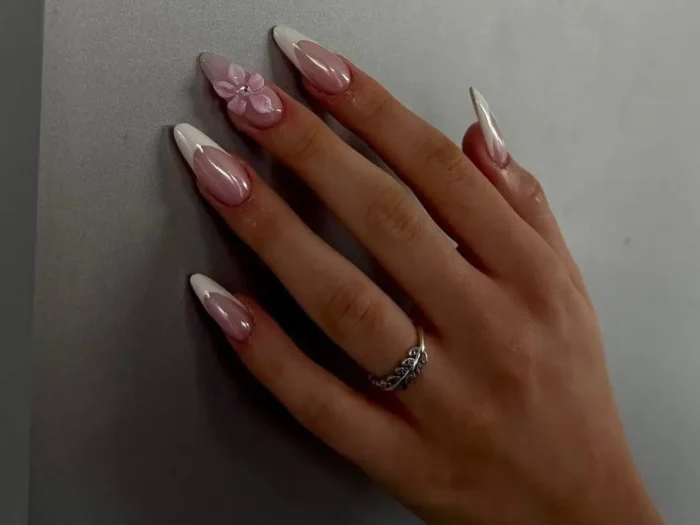 A hand with a classic French manicure on long almond nails, with one nail featuring a 3D dusty pink flower accent.