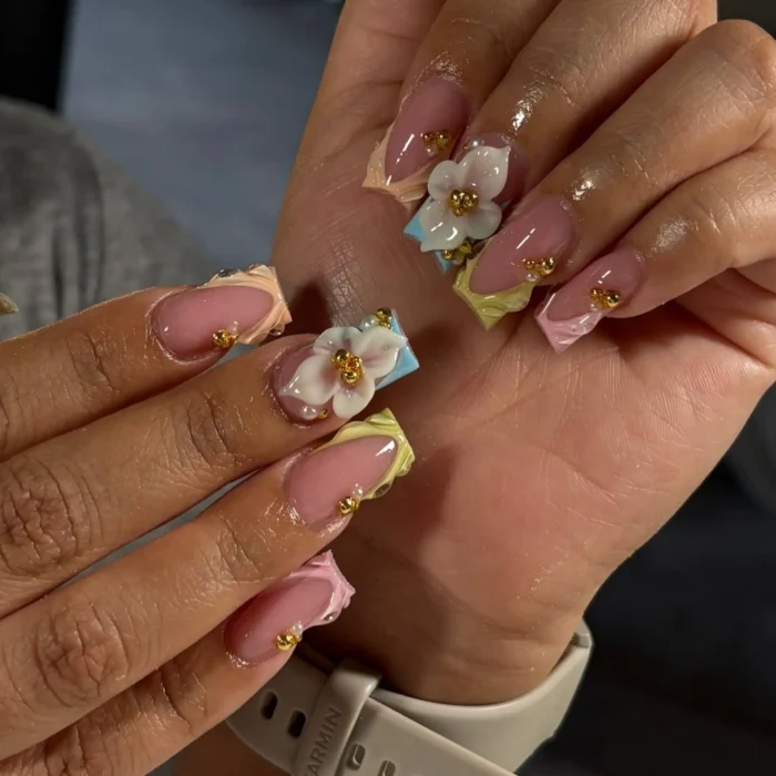 Long coffin-shaped nails displaying textured pastel French tips in yellow, pink, and blue hues, beautifully adorned with white 3D flower charms featuring gold bead centers and delicate gold stud accents.