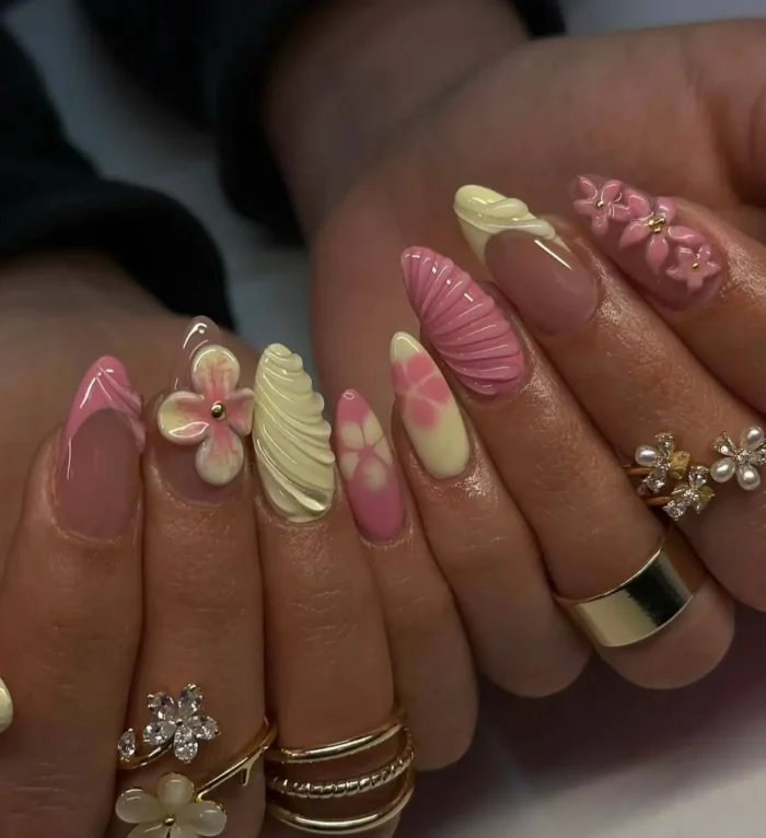 A close-up of a detailed manicure with butter yellow and pink nails, featuring 3D seashell textures and flower art.