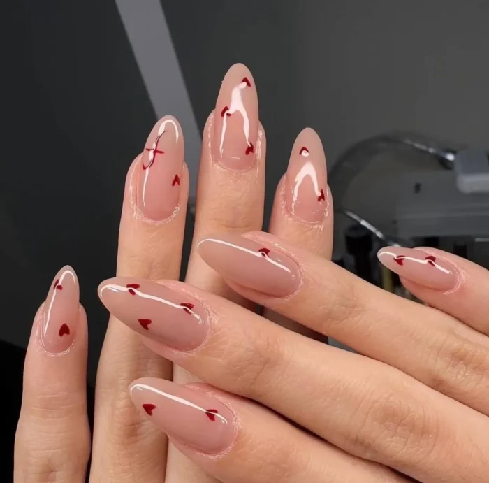 A close-up of long almond-shaped nails with a sheer nude base and tiny red hearts scattered across them for a romantic nail design.