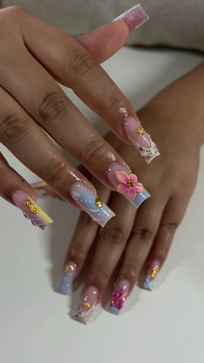 Long square nails showcasing soft pastel ombré tips in blues and yellows, embellished with detailed multi-toned 3D flowers, delicate gold charms, and subtle white dot cloud-like accents.