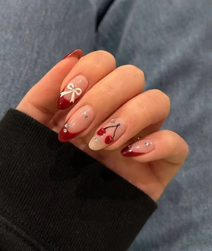 A coquette-style red nail design with deep red French tips, cherry and bow nail art, and rhinestone accents.