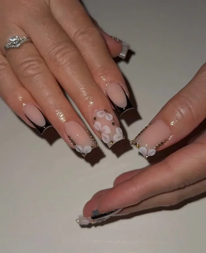 Long square nails displaying a sharp black V-shaped French manicure on a sheer pink base, elegantly embellished with clusters of white 3D flowers and a delicate gold bead outline along the smile line.