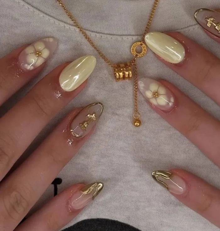 A mismatched butter yellow manicure featuring 3D white flowers, chrome finishes, and abstract molten gold details on almond-shaped nails.