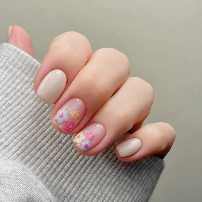 Short, rounded nails with a mix of solid nude polish and sheer nails featuring small, colorful, translucent flower accents.
