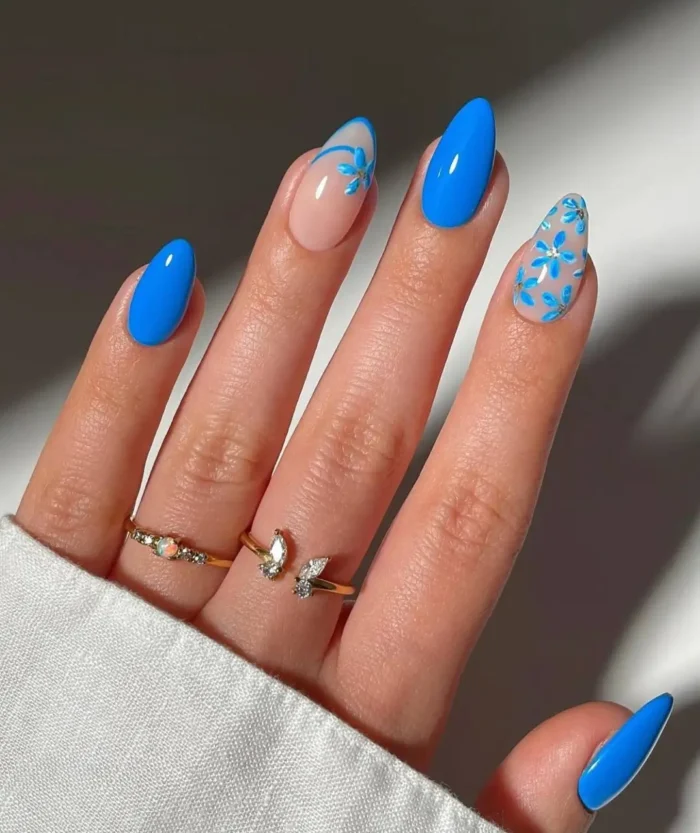 A hand showing a mismatched nail art design with solid cornflower blue nails and accent nails with blue flowers and gold foil.