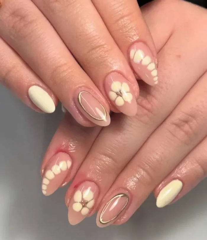A manicure with almond-shaped nails showcasing a mix of solid butter yellow, 3D white and yellow floral art, and gold oval accents on a sheer base.