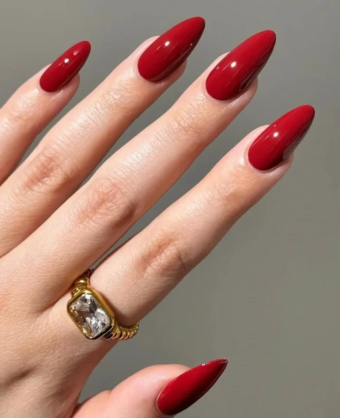 Perfectly manicured medium-long almond-shaped nails painted in a classic, glossy, deep red color, with a gold ring on one finger.