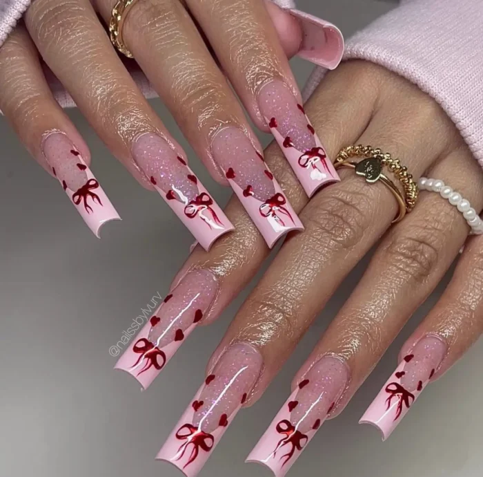 Long square nails with pale pink tips, a sheer shimmer base, tiny floating red hearts, and painted red bow details.