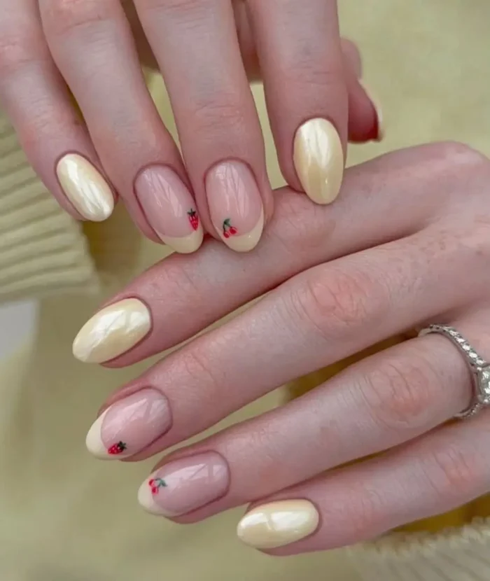 A close-up of oval-shaped nails with a butter yellow manicure featuring cute red strawberry and cherry art alongside pearlescent solid nails.