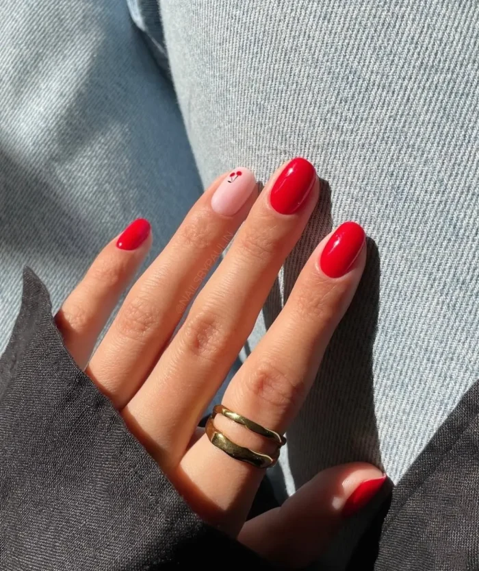 A minimalist red nail design on short nails, with solid red polish and a single accent nail featuring a tiny cherry.