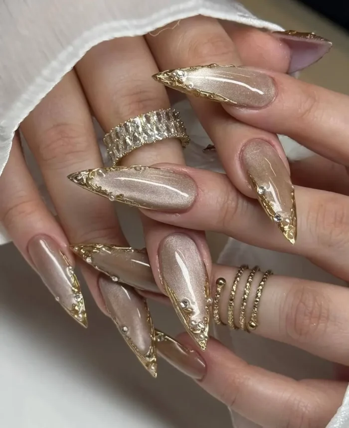 Extra-long stiletto nails with a champagne cat-eye base, framed with textured gold borders and crystal accents for a maximalist nail inspo look.