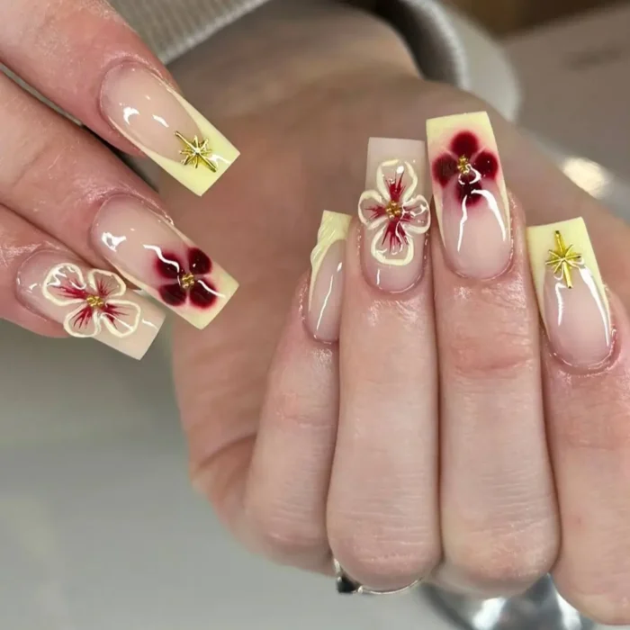 A pale yellow French manicure with accents of 3D flowers that have a deep red center and gold outlines.