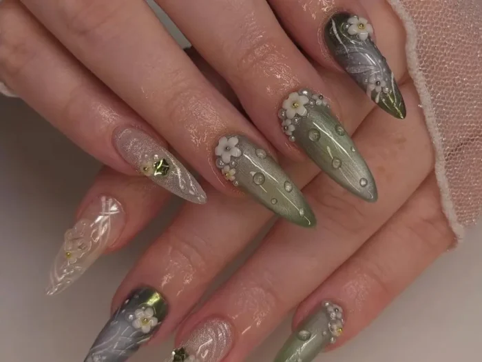 Hand with long stiletto nails featuring beautiful enchanted forest-themed nail art with green velvet shimmer, 3D white flowers, and clear dewdrop effects.