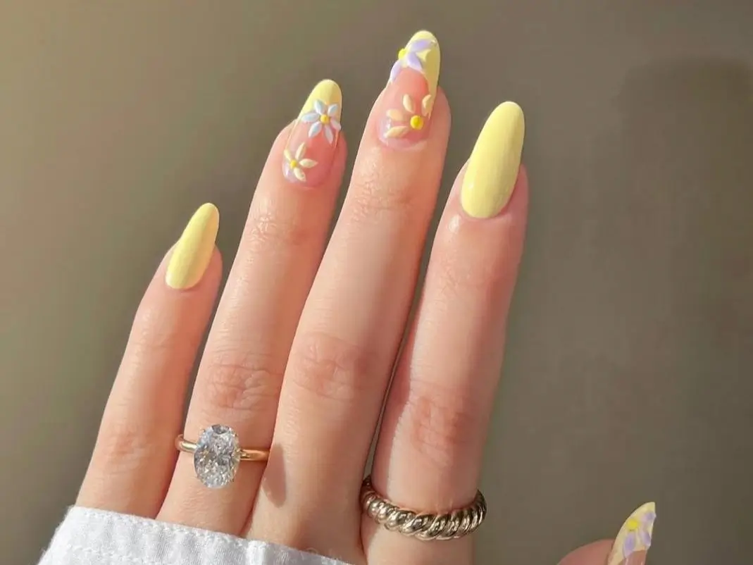 A close-up of a hand with long almond-shaped nails, featuring a chic butter yellow manicure with solid nails and accent nails with small, delicate daisy art.