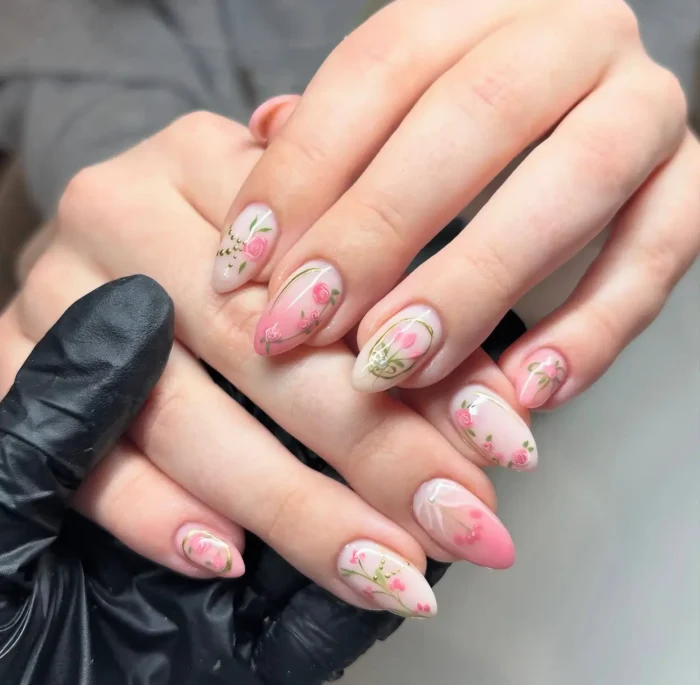 Almond nails featuring a nude to pink gradient base with hand-painted light pink roses, green leaves, and fine gold line accents.