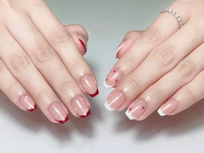 Short squoval nails showing mismatched French manicure: red tips on one hand, white tips with tiny red star accents on the other.