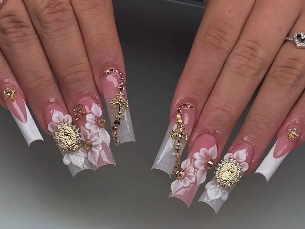 Close-up of extra-long white French tip nails lavishly adorned with 3D white and pink flowers, prominent gold religious icon charms, gold cross charms, and sparkling rhinestone details, showcasing a chic long nail design for 2025.