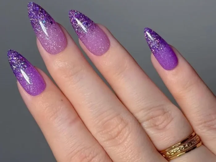 Close-up of elegant almond-shaped nails with a sparkling purple glitter ombré design, fading from dark to light purple.