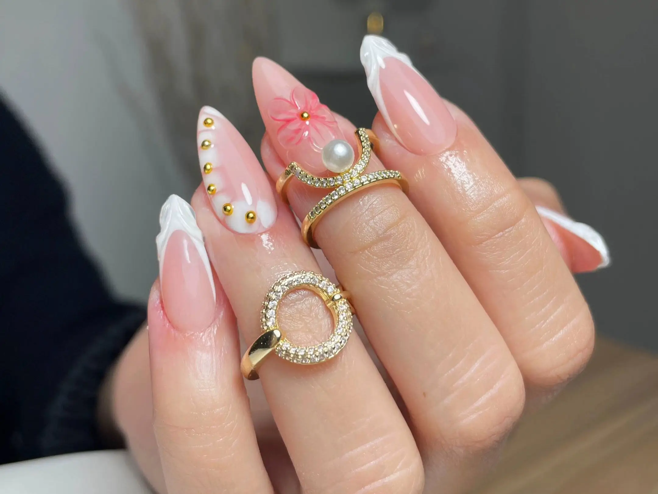 Close-up of long stiletto nails featuring trendy nail art designs: textured white French tips, a 3D pink flower with a pearl center, and gold stud accents, an example from a list of expert picks.