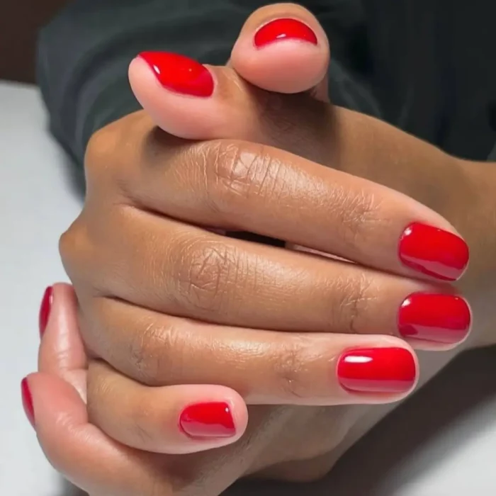 A close-up of dark-toned hands with a classic, glossy cherry red manicure on short, neatly shaped nails.