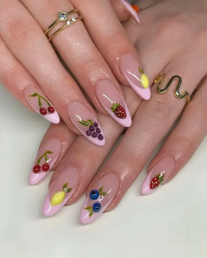 A hand with long almond nails and a chic fruit nail design, featuring mixed fruit art over pink French tips.